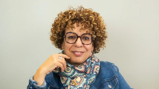 Portrait de Salima Benichou de face. Elle est assise sur un fauteuil noir derrière un mur clair. Elle porte des lunettes a des cheveux bouclés à hauteur d'oreille. Est vêtue d'un pantalon et d'un haut noirs d'une veste en jean et d'un chèche très coloré à motif. Elle sourit sa main droite touche sa joue droite, sa main gauche repose sur sa cuise gauche.